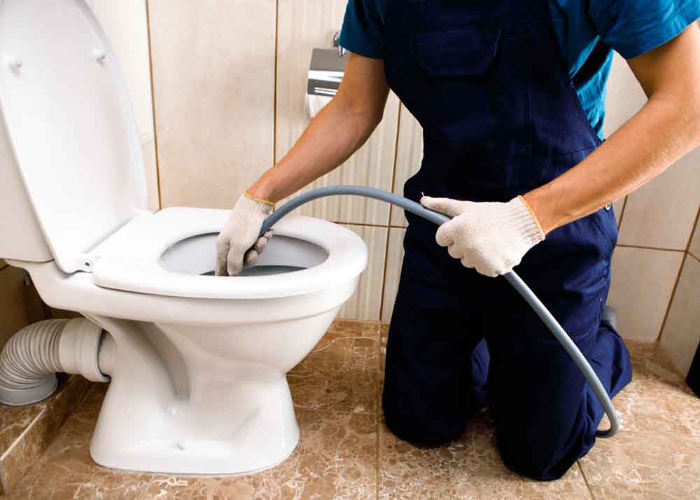 Plumber in navy overalls kneeling on a tiled bathroom floor, using a drain snake tool to unclog a residential toilet