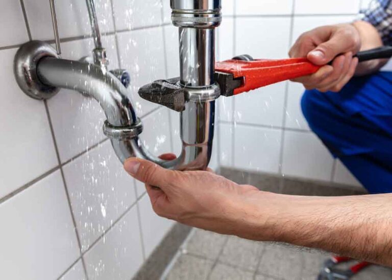 Plumber using a red pipe wrench to tighten a leaking chrome drainpipe under a tiled sink with water spraying from the joint