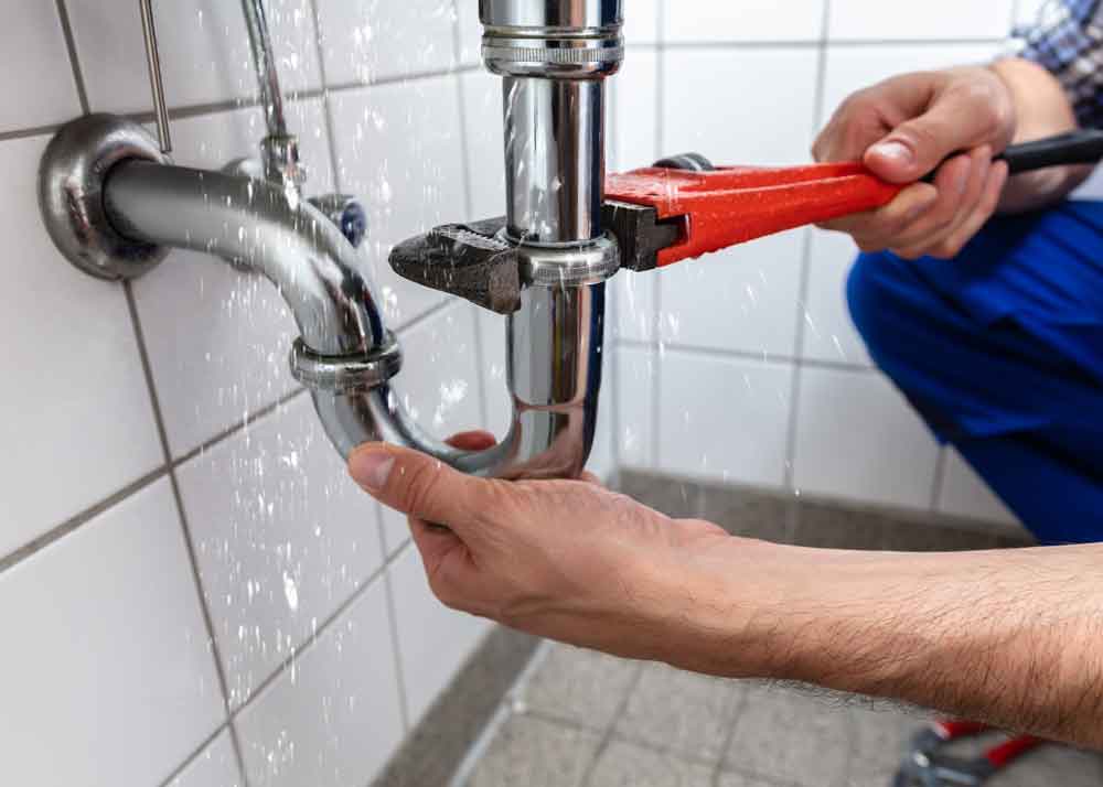 Plumber using a red pipe wrench to tighten a leaking chrome drainpipe under a tiled sink with water spraying from the joint