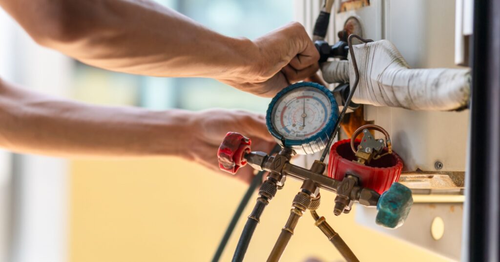 Technician performing AC maintenance using a manifold gauge to check refrigerant pressure on an outdoor air conditioning unit.
