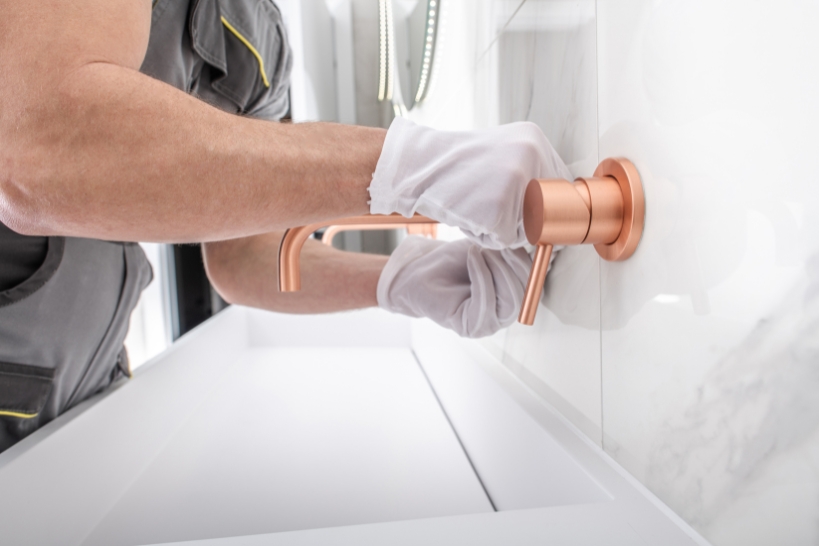 Plumber installing a wall-mounted copper faucet above a modern bathroom sink.
