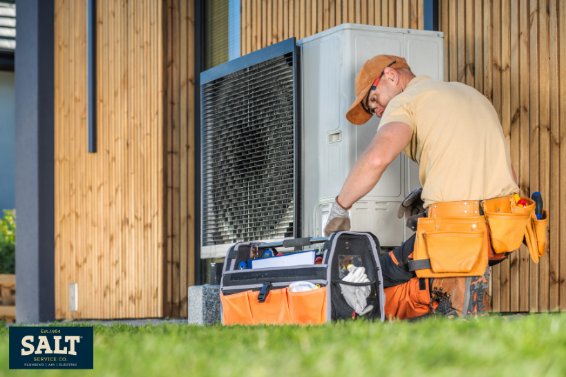 HVAC technician performing seasonal spring maintenance on an outdoor air conditioning unit, with tools and service bag placed beside the condenser.