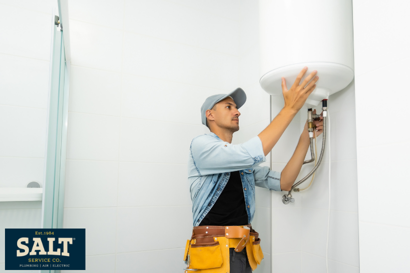 Plumber installing or servicing a wall-mounted water heater in a bathroom, wearing a tool belt and cap during maintenance.