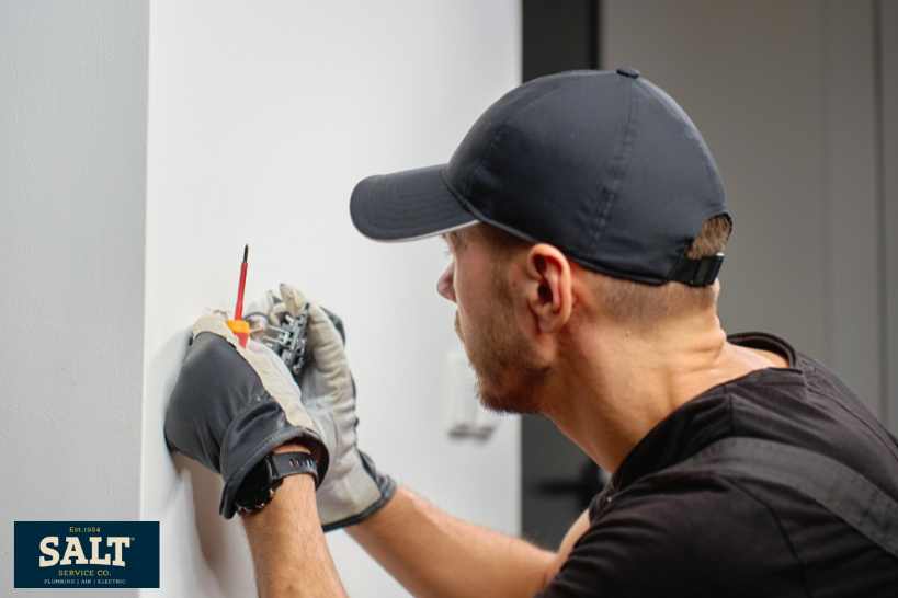 Electrician wearing a cap and work gloves installs or repairs an electrical outlet on an interior wall using a screwdriver and hand tools.