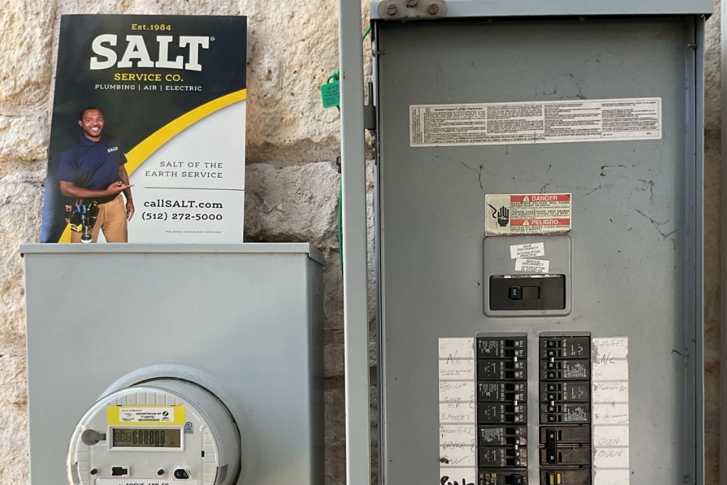 Electrical meter and breaker panel with service company card, labeled circuits, and safety warnings during residential electrical inspection or repair